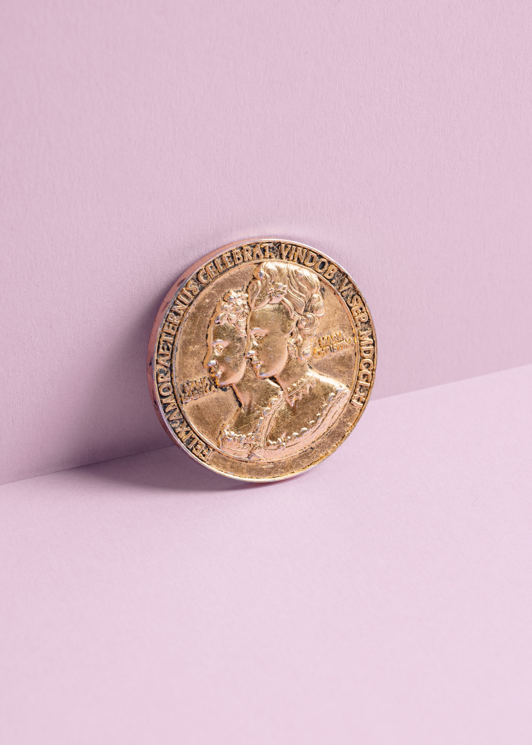 Gold commemorative coin featuring two profiles against a pastel pink background.