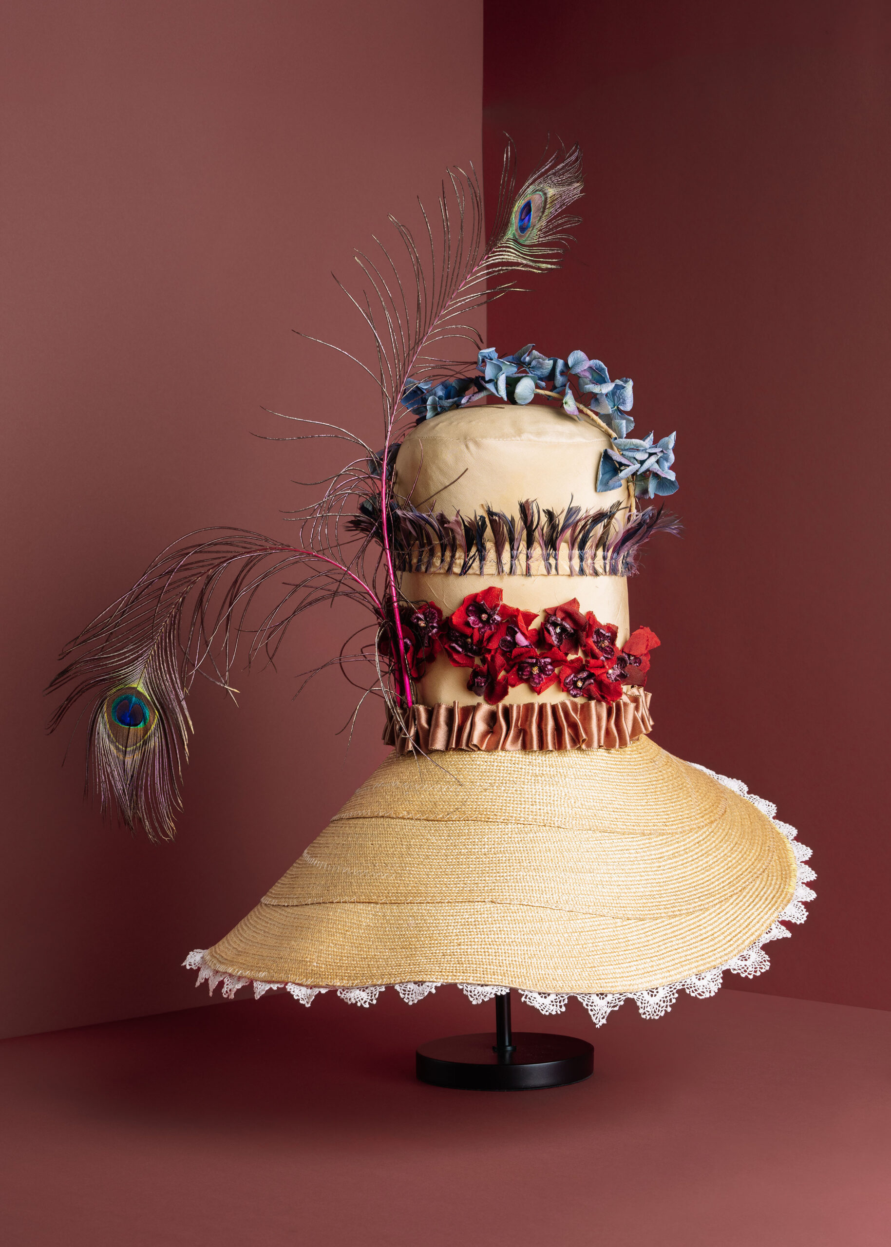 Elegant straw hat adorned with colorful flowers and peacock feathers, displayed on a stand.