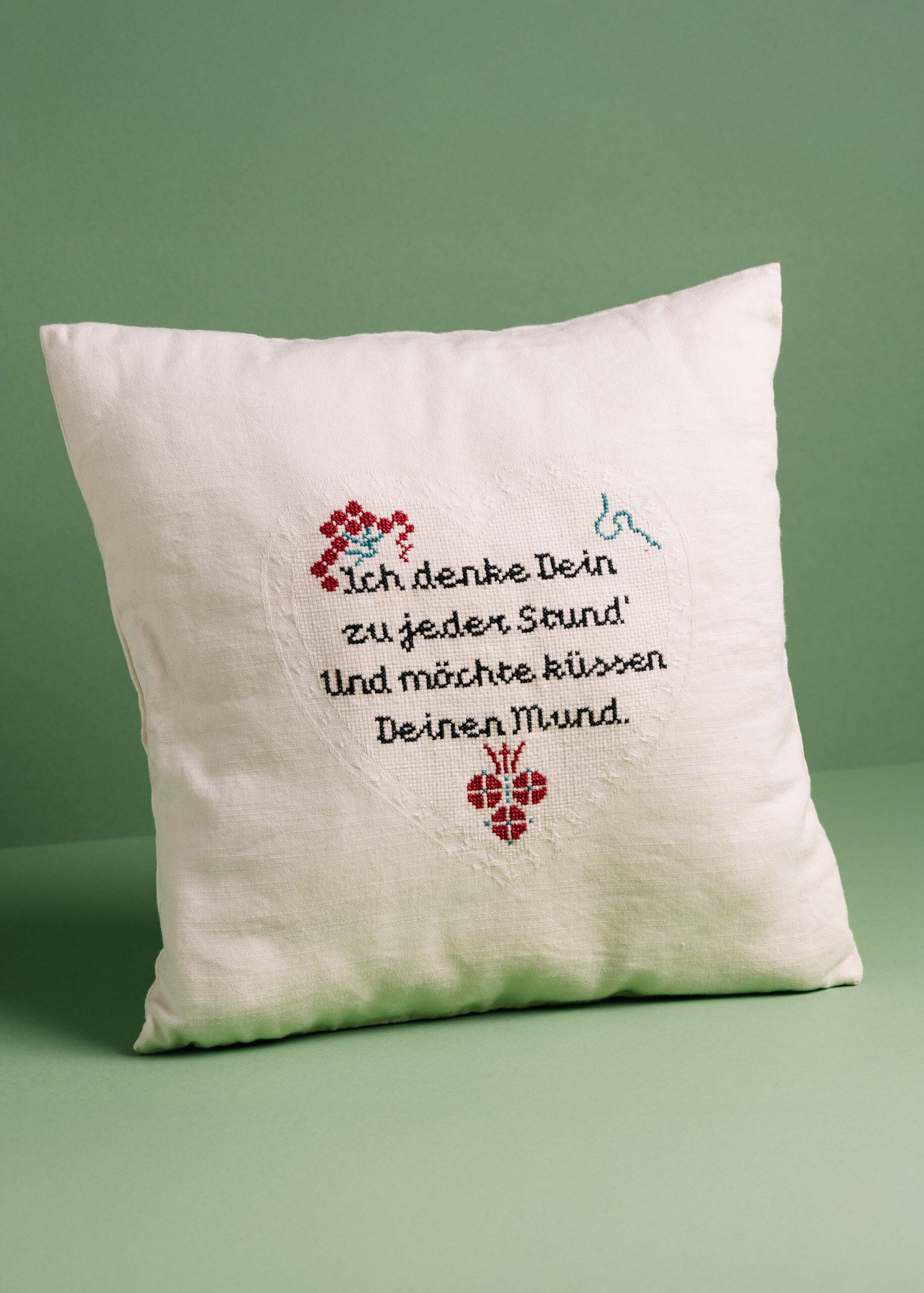 Embroidered pillow with romantic German text and decorative hearts on a green background.
