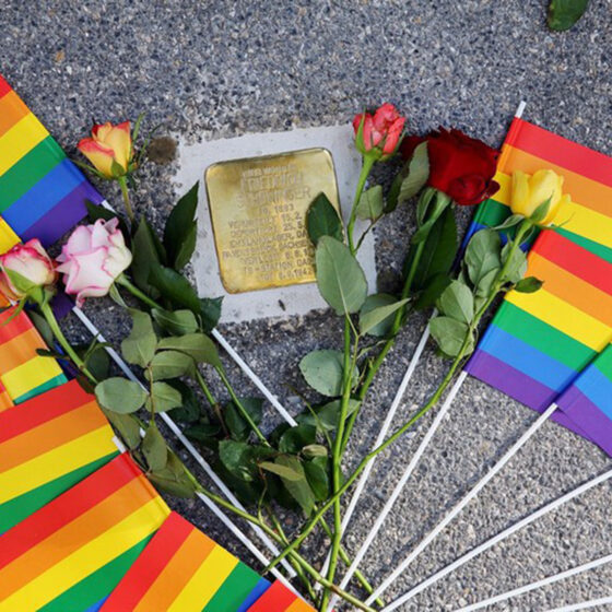 Regenbogenflaggen und Rosen umrahmen eine Gedenktafel für LGBTQ+-Rechte.