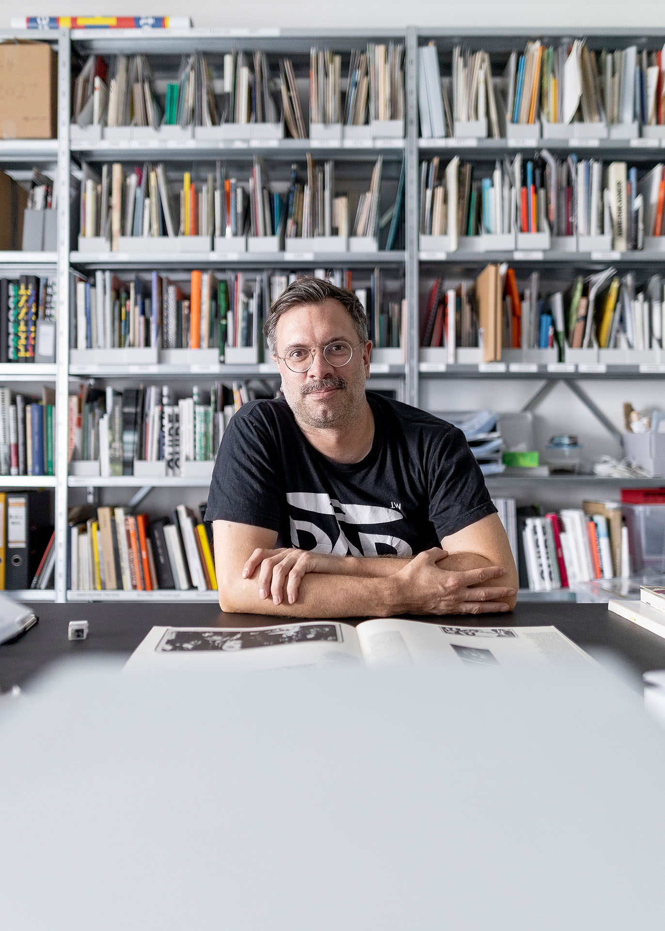 Person sitting at a desk with books, looking directly at the camera in a studio.
