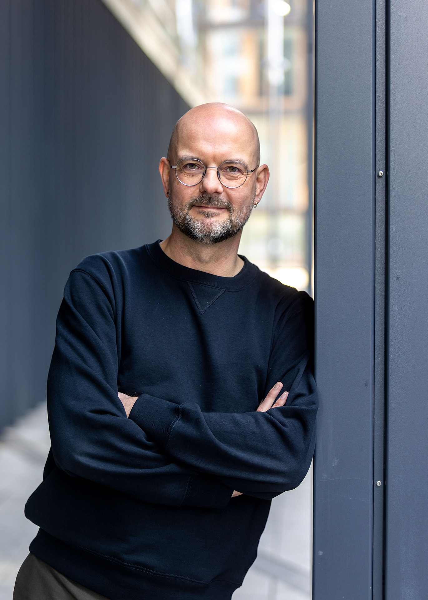 Person with glasses and a beard, leaning against a wall, arms crossed.