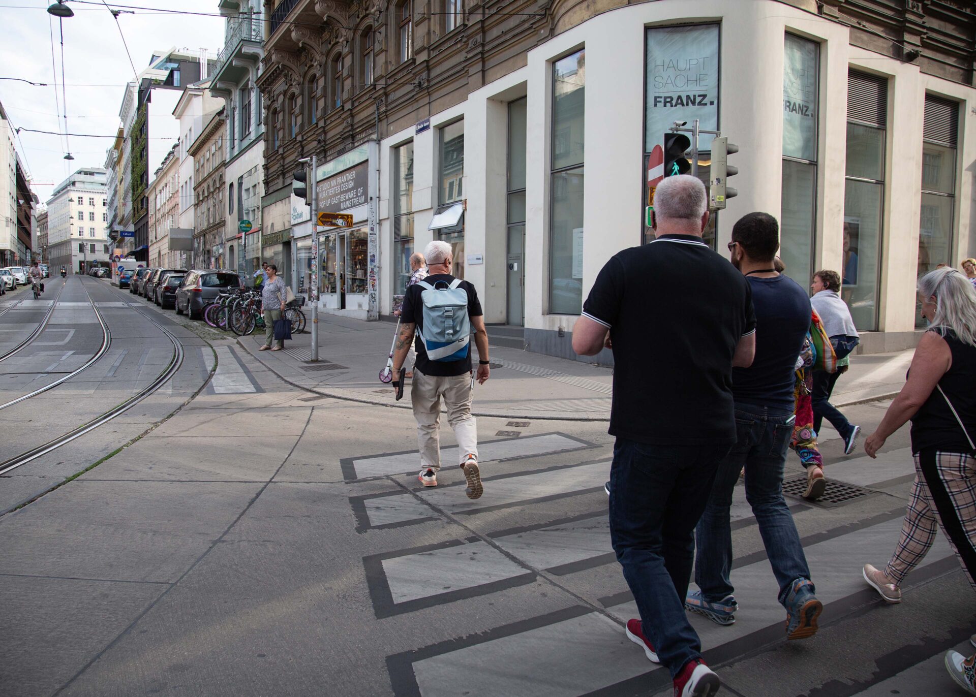 Pedestrians crossing a street near shops and tram tracks in an urban area.