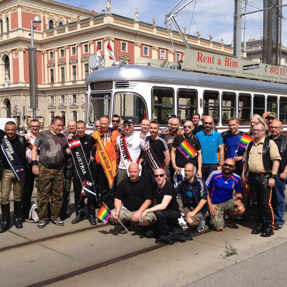 Gruppe von Menschen mit Regenbogenflaggen vor einer Straßenbahn in Wien, feiernd und lächelnd.