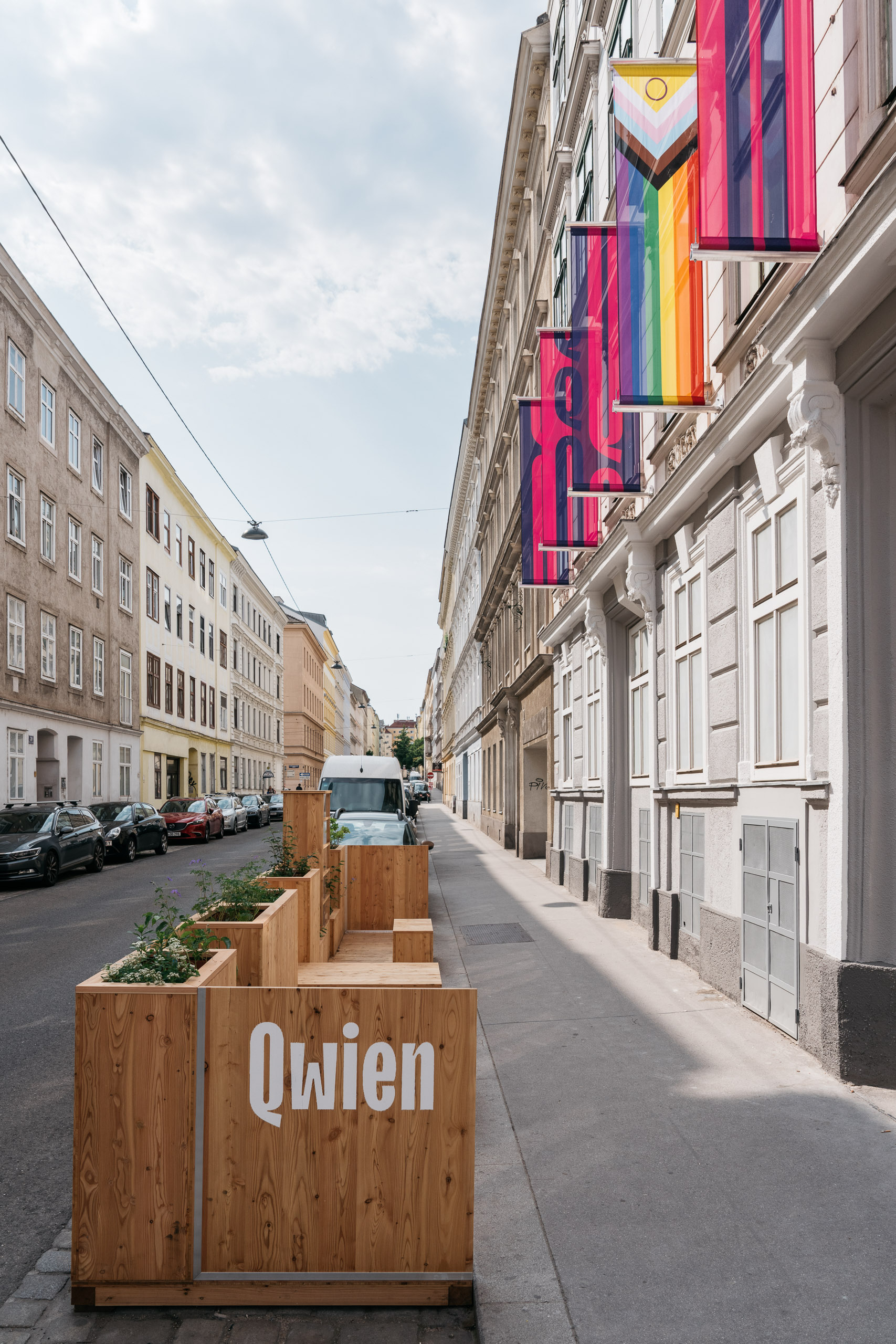 Holzbank mit "Qwien"-Schriftzug auf belebter Straße, Regenbogenflaggen im Hintergrund.