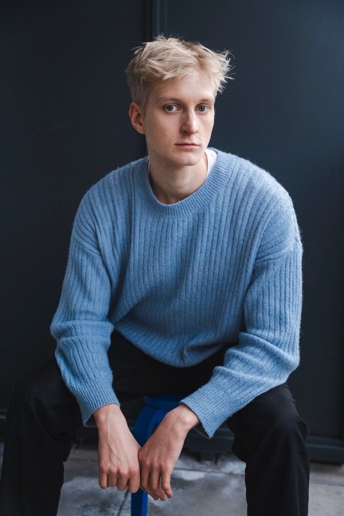 Young person in a blue sweater sitting on a stool, gazing thoughtfully.