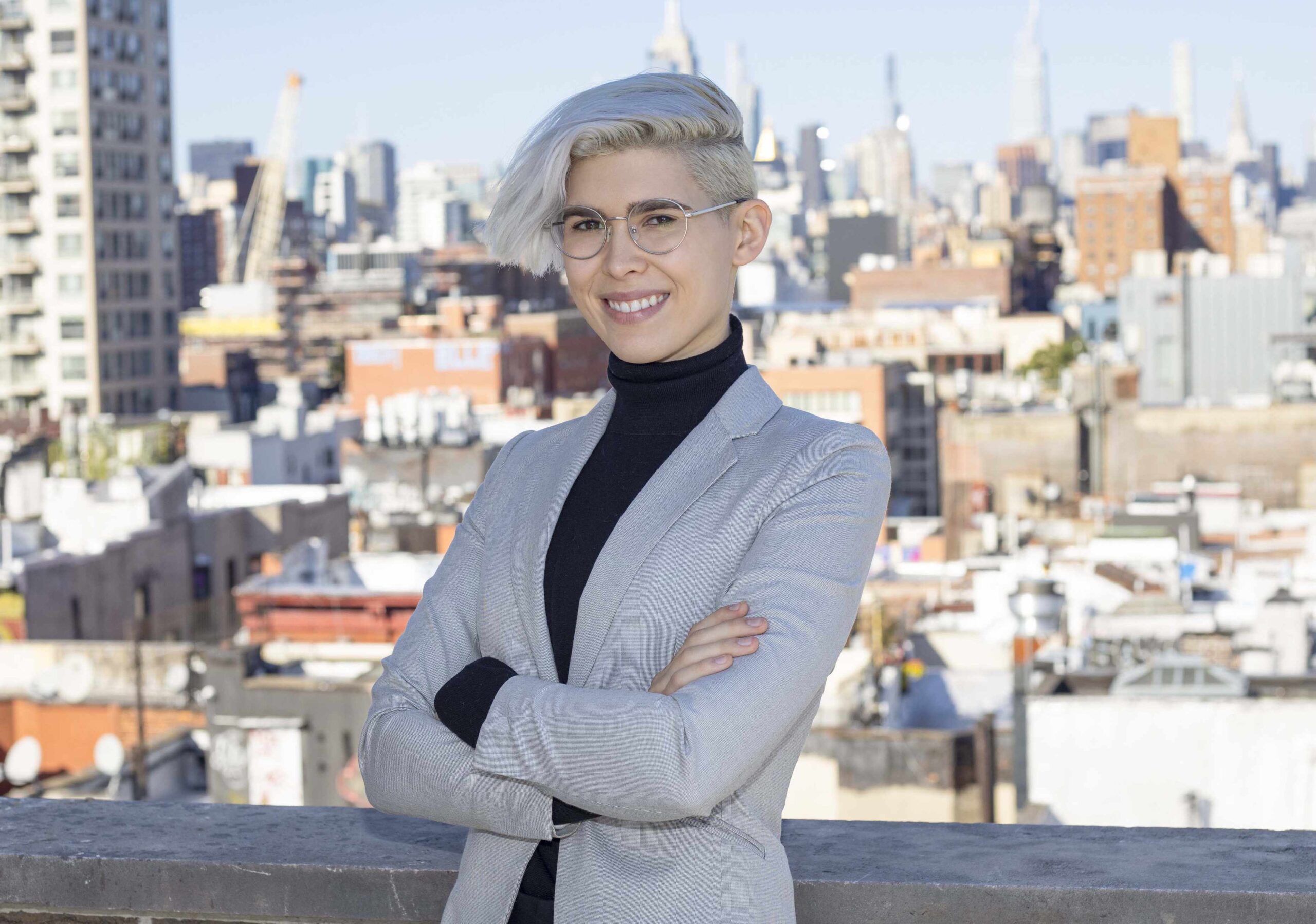 Confident individual in a suit poses with crossed arms against a city skyline backdrop.